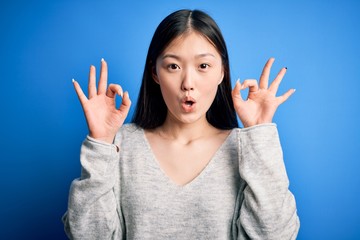 Wall Mural - Young beautiful asian woman wearing casual sweater standing over blue isolated background looking surprised and shocked doing ok approval symbol with fingers. Crazy expression