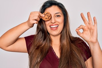 Young hispanic woman eating sweet chocolated chips cookie over isolated background doing ok sign with fingers, excellent symbol