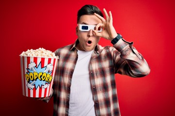 Wall Mural - Young handsome caucasian man wearing 3d movie glasses and eating popcorn doing ok gesture shocked with surprised face, eye looking through fingers. Unbelieving expression.