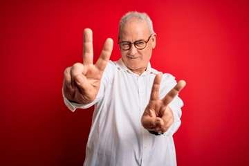 Middle age handsome hoary man wearing casual shirt and glasses over red background smiling with tongue out showing fingers of both hands doing victory sign. Number two.