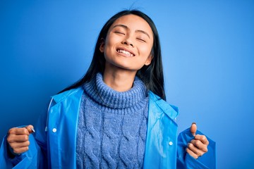 Wall Mural - Young beautiful chinese woman wearing rain coat standing over isolated blue background very happy and excited doing winner gesture with arms raised, smiling and screaming for success. Celebration