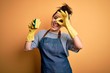 Young brunette cleaner woman wearing housekeeping gloves holding scourer scrub with happy face smiling doing ok sign with hand on eye looking through fingers