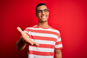Wall Mural - Young handsome african american man wearing casual striped t-shirt and glasses smiling cheerful offering palm hand giving assistance and acceptance.