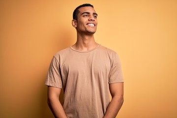 Poster - Young handsome african american man wearing casual t-shirt standing over yellow background looking away to side with smile on face, natural expression. Laughing confident.
