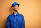 Fototapeta  - Young handsome african american worker man wearing blue uniform and security helmet looking away to side with smile on face, natural expression. Laughing confident.