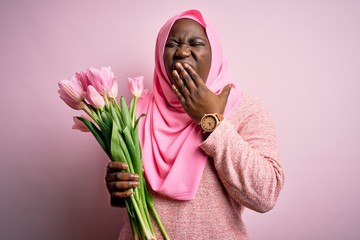 Poster - Young african american plus size woman wearing muslim hijab holding bouquet of pink tulips bored yawning tired covering mouth with hand. Restless and sleepiness.