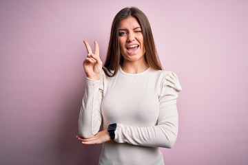 Young beautiful woman with blue eyes wearing casual white t-shirt over pink background smiling with happy face winking at the camera doing victory sign with fingers. Number two.