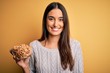 © Krakenimages.com - Young beautiful brunette woman holding bowl with peanuts over isolated yellow background with a happy face standing and smiling with a confident smile showing teeth