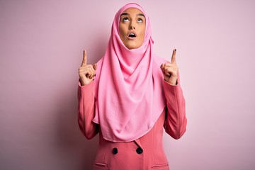 Young beautiful brunette businesswoman wearing pink muslim hijab and business jacket amazed and surprised looking up and pointing with fingers and raised arms.