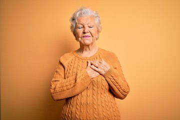 Senior beautiful grey-haired woman wearing casual sweater over isolated yellow background smiling with hands on chest with closed eyes and grateful gesture on face. Health concept.