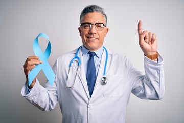 Sticker - Middle age handsome grey-haired doctor man wearing coat holding blue cancer ribbon surprised with an idea or question pointing finger with happy face, number one