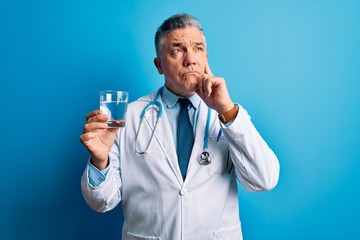 Poster - Middle age handsome grey-haired doctor man drinking glass of water over blue background serious face thinking about question, very confused idea