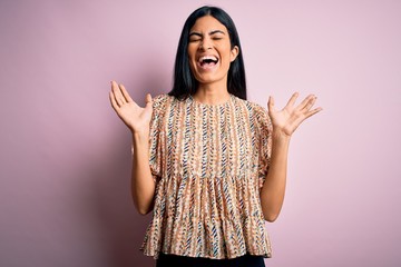 Young beautiful hispanic fashion woman wearing elegant style over pink background celebrating mad and crazy for success with arms raised and closed eyes screaming excited. Winner concept