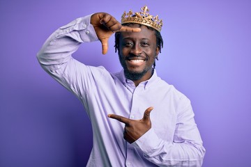 Sticker - Young african american man wearing golden crown of king over isolated purple background smiling making frame with hands and fingers with happy face. Creativity and photography concept.