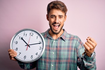 Young man with beard doing countdown using big clock over isolated pink background screaming proud and celebrating victory and success very excited, cheering emotion