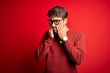 © Krakenimages.com - Young handsome man with beard wearing glasses and sweater standing over red background rubbing eyes for fatigue and headache, sleepy and tired expression. Vision problem