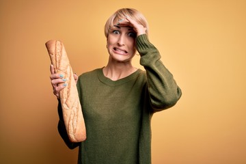 Poster - Young blonde woman with short hair holding fresh bread baguette over yellow background stressed with hand on head, shocked with shame and surprise face, angry and frustrated. Fear and upset