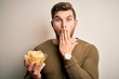 © Krakenimages.com - Young blond man with beard and blue eyes holding bowl with unhealthy potatoes chips cover mouth with hand shocked with shame for mistake, expression of fear, scared in silence, secret concept