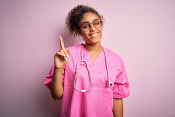 Poster - African american nurse girl wearing medical uniform and stethoscope over pink background showing and pointing up with finger number one while smiling confident and happy.