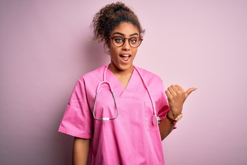 Poster - African american nurse girl wearing medical uniform and stethoscope over pink background smiling with happy face looking and pointing to the side with thumb up.