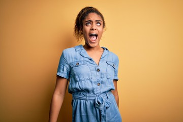 Sticker - Young beautiful african american girl wearing denim dress standing over yellow background angry and mad screaming frustrated and furious, shouting with anger. Rage and aggressive concept.