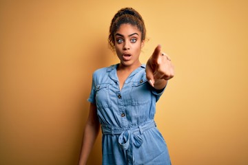 Sticker - Young beautiful african american girl wearing denim dress standing over yellow background Pointing with finger surprised ahead, open mouth amazed expression, something on the front