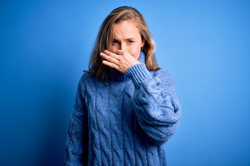 Poster - Young beautiful blonde woman wearing casual turtleneck sweater over blue background smelling something stinky and disgusting, intolerable smell, holding breath with fingers on nose. Bad smell