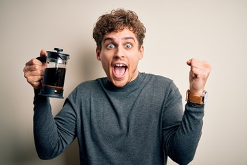 Young blond man with curly hair making coffee using coffemaker over white background screaming proud and celebrating victory and success very excited, cheering emotion