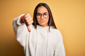 Canvas Print - Young beautiful asian woman wearing casual sweater and glasses over yellow background looking unhappy and angry showing rejection and negative with thumbs down gesture. Bad expression.