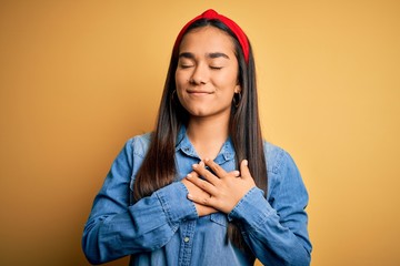 Canvas Print - Young beautiful asian woman wearing casual denim shirt and diadem over yellow background smiling with hands on chest with closed eyes and grateful gesture on face. Health concept.