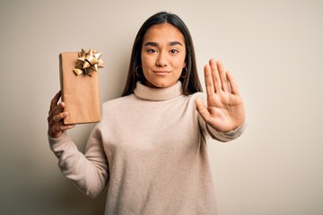 Wall Mural - Young beautiful asian woman holding birthday gift standing over isolated white background with open hand doing stop sign with serious and confident expression, defense gesture