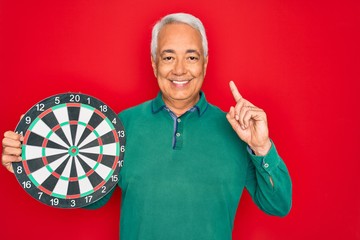 Wall Mural - Middle age senior grey-haired man holding competition dartboard target over red background surprised with an idea or question pointing finger with happy face, number one