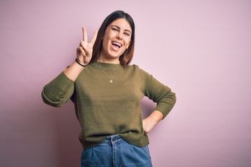 Sticker - Young beautiful woman wearing casual sweater standing over isolated pink background smiling with happy face winking at the camera doing victory sign. Number two.