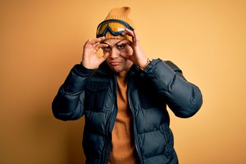 Wall Mural - Young brazilian skier man wearing snow sportswear and ski goggles over yellow background Trying to open eyes with fingers, sleepy and tired for morning fatigue