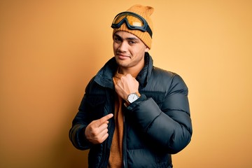 Wall Mural - Young brazilian skier man wearing snow sportswear and ski goggles over yellow background In hurry pointing to watch time, impatience, looking at the camera with relaxed expression