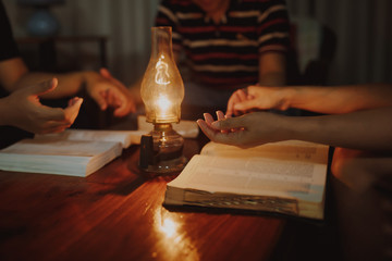 Christian small group  holding hands and raise a hand while praying together over blurred bible  with a vintage lamp on wooden table in the home at night, Christian home worship  concept