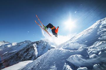 Wall Mural - Low angle view athlete skier in an orange jacket does a back flip with flying powder of snow against a clear blue sky sun and snow-capped mountains of the Caucasus.