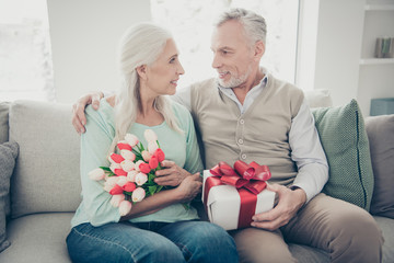 Poster - Adorable aged pair sitting comfy sofa celebrating anniversary hold hands red tulips bunch and giftbox