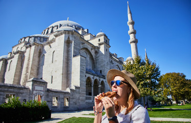 Wall Mural - Happy attractive woman tourist in hat, eating Simitci (a round bagel with sesame seeds) against the background of Suleymaniye mosque, Istanbul,Turkey. Religion and travel concept.