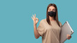 © South House Studio - Cute woman in medical mask shows ok sign, she holds her laptop computer isolated over blue