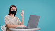 © South House Studio - Pretty brunette female works with her computer isolated, she shows ok sign and wear medical mask, isolated over blue bg