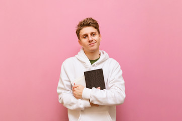 Wall Mural - Portrait of handsome student boy with blond hair and in white hoodie stands on pink background and poses at camera with books in hands. Young blond man holding book in hand isolated on pink background