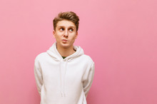 Funny Guy In White Hoodie Stands On A Pink Background With A Smile On His Face And Looks Away At Copy Space. Cheerful Young Man In Casual Clothes Isolated On Pink Background. Copy Space