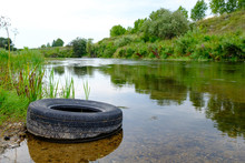 The Tire From The Car Lies On The Bank Of The River Polluting The Nature