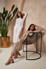 Wall Mural - Two beautiful sexy brunette woman friends face cosmetic makeup tanned skin wear fashion clothes style beige white knitted suit accessory interior furniture armchair journey summer flowerpot palm boho.