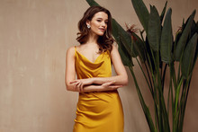 Portrait Of Beautiful Woman Fashion Model Brunette Hair Wear Yellow Silk Dress Accessory Jewelry Earrings Cosmetic Face Clothes Romantic Date Party Walk Summer Collection Tanned Skin Green Leaves.