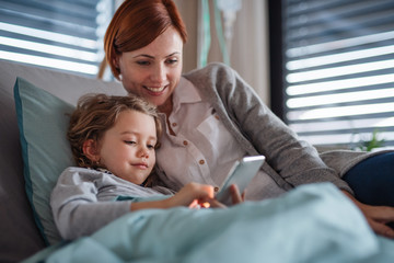 Wall Mural - Small girl with mother in bed in hospital, using smartphone to pass time.