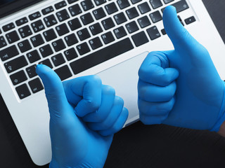 Two hands in blue gloves showing thumbs up, Portable laptop. Coronavirus protection.