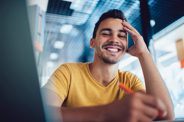 Smiling handsome male student enjoying learning process making notes for homework,portrait of successful smart man journalist satisfied with job writing article in cafe interior for publication.