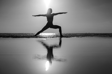 Silhouette of woman standing at yoga pose on the tropical beach during sunset. Girl practicing yoga near sea water. Black and white